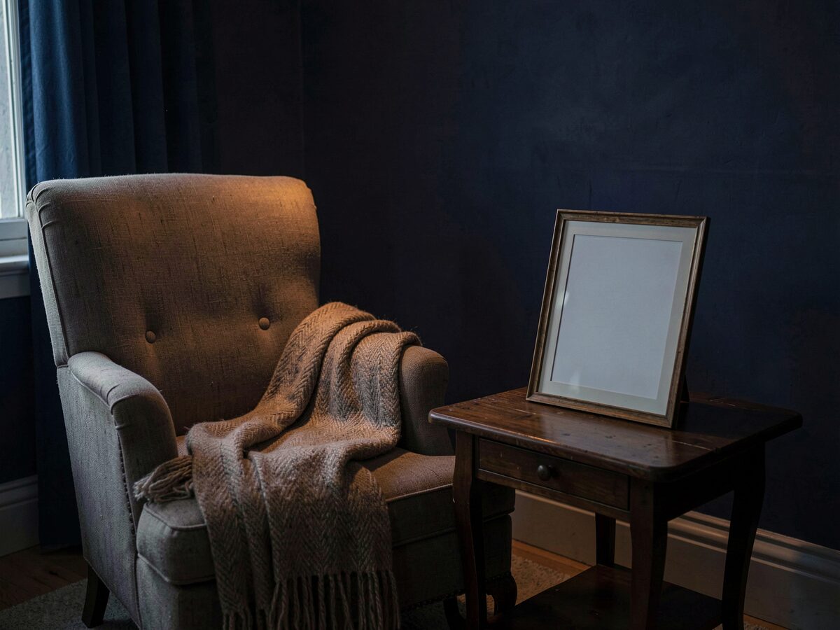 an empty armchair with a folded shawl beside a small table holding a framed blank photograph turned face down, dim warm light, no people, no readable text