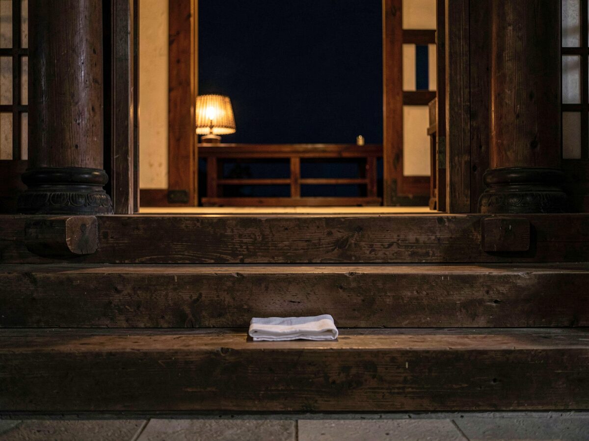 three wooden steps in warm shadow leading up to a low lit doorway, a folded white cloth on the first step, quiet ceremonial stillness, no people, no text